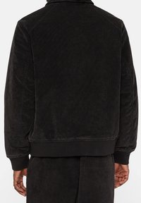 Czarna bawełniana kurtka o miękkiej, teksturowanej powierzchni, z klasycznym kołnierzem i prążkowanymi mankietami. Prosty krój bez widocznych wzorów czy elementów metalowych.