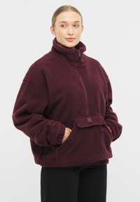 Burgundy színű polár pulóver magas gallérral, cipzáras záródással, elülső zsebbe és elasztikus derékrésszel. Puha tapintás és laza szabás.