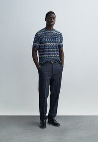 Cămașă cu mâneci scurte cu model zigzag în tonuri de albastru și verde, asortată cu pantaloni închiși la culoare, texturizați și pantofi negri.