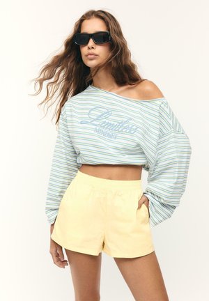 Junge Frau trägt ein übergroßes, gestreiftes Crop-Top, das schulterfrei ist, gelbe Shorts und große schwarze Sonnenbrillen, posiert mit einer Hand in der Tasche.