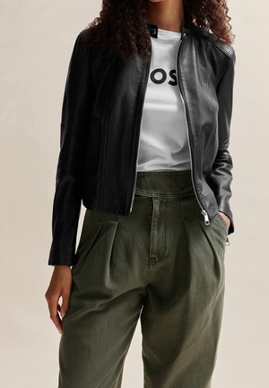 Personne portant une veste en cuir noire sur une chemise blanche avec un texte noir et un pantalon plissé vert olive taille haute avec les mains dans les poches.