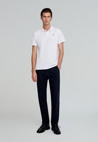 Camisa polo blanca de manga corta con cuello y un pequeño logo en el pecho. Combinada con pantalones azul marino oscuros y zapatos negros.