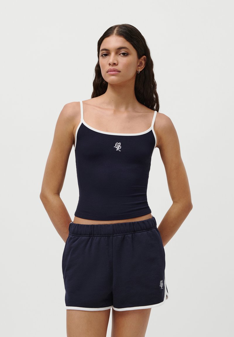 Top corto blu con rifiniture bianche e logo, abbinato a pantaloni corti blu con accenti bianchi. Entrambi sono realizzati in un tessuto morbido e elastico.