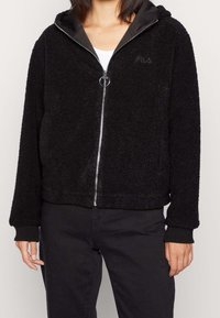 Czarna, puszysta bluza z kapturem zapinanym na zamek, z podszytym kapturem, ściągaczami przy mankietach i srebrnym zamkiem błyskawicznym. Na piersi znajduje się subtelne logo, a powierzchnia ma teksturowaną strukturę.