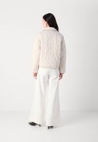 Jachetă bej matlasată cu guler înalt, cu cusături în formă de diamant și mâneci lungi, asortată cu pantaloni albi evazați și pantofi cu toc negri.