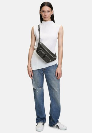Borsa a tracolla in pelle nera con tre tasche con zip, indossata sopra una canotta bianca testurizzata e jeans blu strappati.