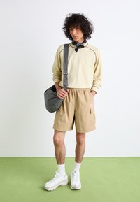 Light beige long-sleeve shirt with decorative stitching, tan shorts, white sneakers, and a checkered shoulder bag. Textured, casual design.