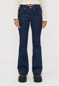 Dark blue flared jeans made of denim, featuring front pockets, a button closure, and contrasting stitching. Worn with black platform shoes.