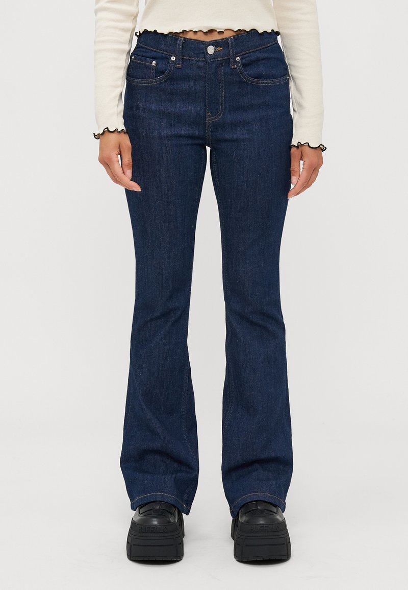 Dark blue flared jeans made of denim, featuring front pockets, a button closure, and contrasting stitching. Worn with black platform shoes.
