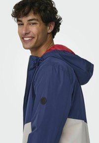 Marineblauwe en witte kleurenblokjas met een rode capuchon, gemaakt van gladde stof. Voorzien van een ritssluiting aan de voorkant en een logopatch op de linkermouw.