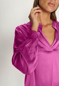Blouse en satin rose vif avec un col en V, manches longues agrémentées de boutons, et une texture lisse et brillante.