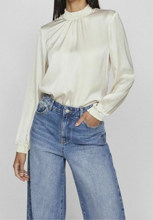Femme portant un chemisier en satin crème avec col et poignets smockés, associé à un jean taille haute, jambes larges, bleu clair.