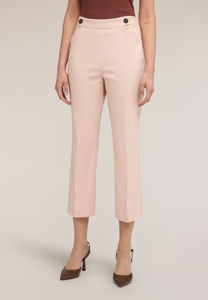 Frau trägt hellrosa, knöchellange, maßgeschneiderte Hose mit vorderen Knöpfen, kombiniert mit braunen, spitzen Schuhen mit Absatz und einem ärmellosen Oberteil.