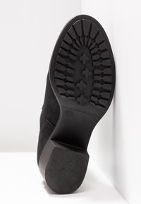 Suela de bota negra con profundas marcas y patrones en forma de cruz, tamaño 37, inclinada contra un fondo de pared y suelo blancos.