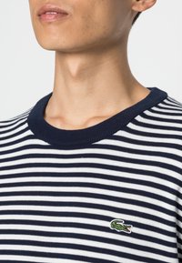 Man wearing navy and white striped shirt with green crocodile logo on the chest, shown from neck to mid-torso against white background.