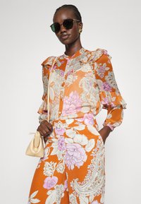 Blusa floreale colorata in arancione e viola, con maniche con volant e chiusura a bottoni. Abbinata a pantaloni coordinati con motivo e una piccola borsa beige.