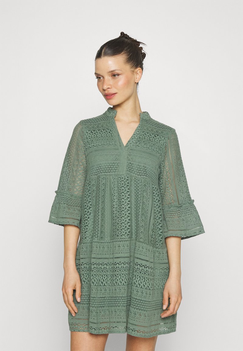 Rochie verde din dantelă, cu decolteu în formă de V, mâneci trei sferturi și modele intricate. Material texturat, ușor, cu elemente de design stratificate.