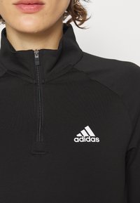Schwarzes sportliches Halbzipp-Oberteil mit hohem Kragen, glatter Textur und einem weißen Adidas-Logo auf der linken Brustseite.