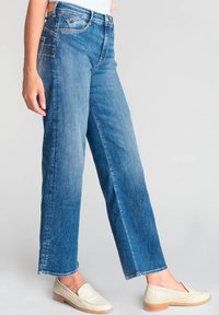 Jeans bleu clair taille haute avec une coupe décontractée, comportant cinq poches et une texture lisse. Portés avec des mocassins de couleur claire.