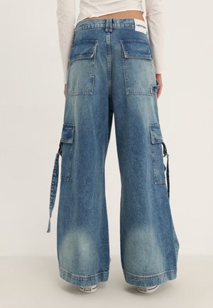 Person trägt blaue Denim-Cargohosen mit weitem Bein, mehreren Taschen hinten und an den Seiten und einem weißen Aufnäher an der Taille.