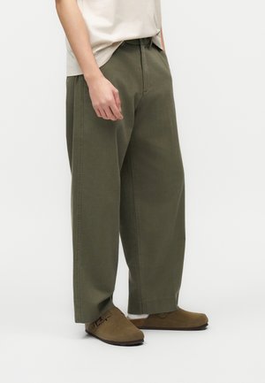 Oseba, oblečena v široke hlače olivno zelene barve, bež zgornji del, bele nogavice in rjave semiš mule, stoji na enobarvnem ozadju.