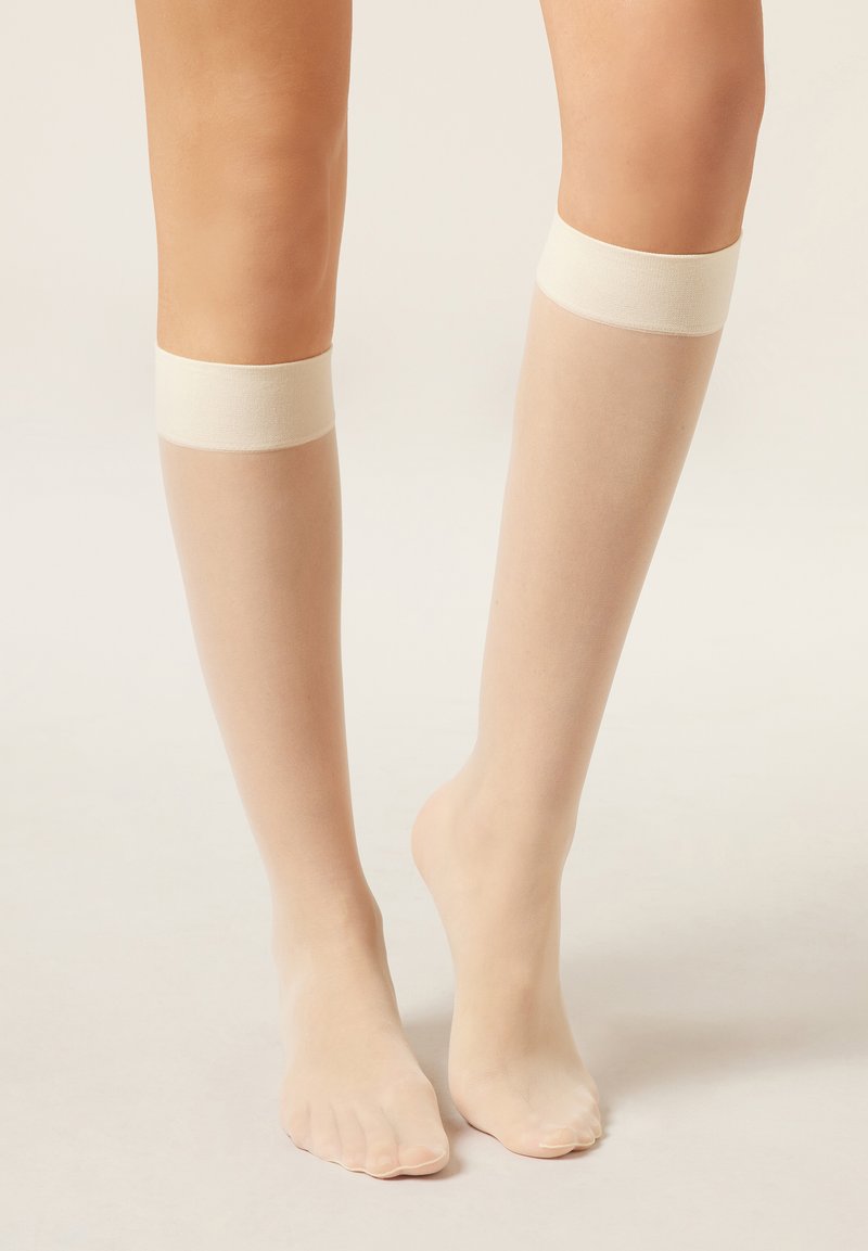 Calze a rete nude trasparenti fino al ginocchio con una texture liscia, caratterizzate da fasce elastiche color crema nella parte superiore. Le dita dei piedi sono visibili e il design è minimalista.
