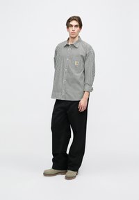 Camisa de botones a rayas en blanco y negro, con un bolsillo en el pecho que presenta un logo. Combinada con pantalones anchos negros y calzado gris.