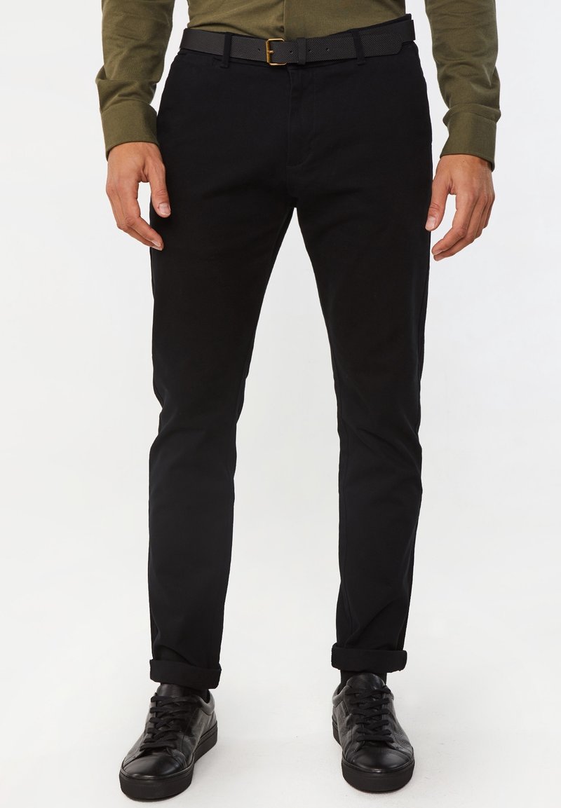 Black cotton trousers with a slim fit, featuring a rolled cuff, side pockets, and a black belt with a gold buckle. Paired with black sneakers.