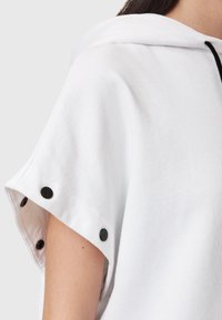 Sudadera blanca con capucha y mangas cortas, que presenta botones a presión negros en los bordes de las mangas. Tejido suave con una textura lisa.