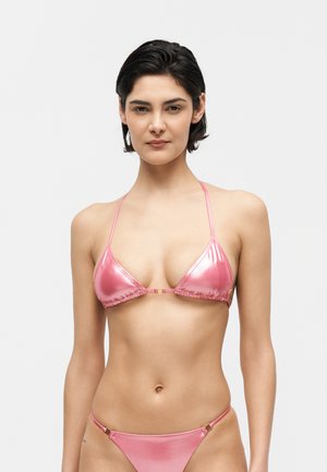 Kobieta z krótkimi ciemnymi włosami, ubrana w błyszczący różowy trójkątny top bikini i pasujący dół na jednolitym białym tle.