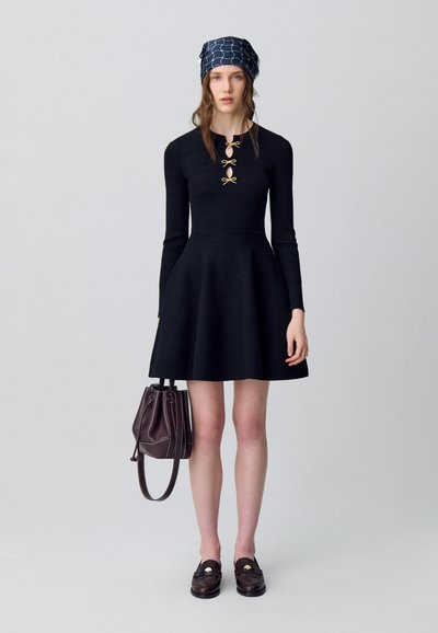 Donna che indossa un vestito lungo e nero con dettagli a fiocco, un foulard blu con motivi, tiene una borsa marrone e indossa mocassini marroni scuri.