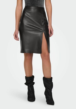 Femme portant une jupe en cuir noir jusqu'aux genoux avec une fente à l'avant, associée à des bottes à talons noires décontractées, se tenant sur un fond blanc.