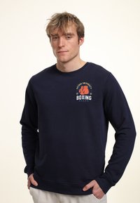 Felpa di colore blu navy realizzata in tessuto morbido, con scollo rotondo e maniche lunghe. La parte frontale presenta una stampa grafica con guantoni da boxe e testo.