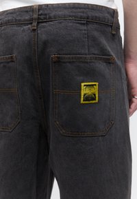 Zadný pohľad na osobu nosiacu čierne rifle z denimu s oranžovým prešívaním a žltou obdĺžnikovou náplasťou na pravom zadnom vrecku.