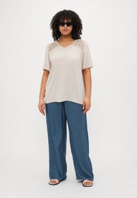 Beige kortermet bluse med heklete accenter på skuldrene, kombinert med løse blå bukser og flate sandaler. Glatt, lett stoff.