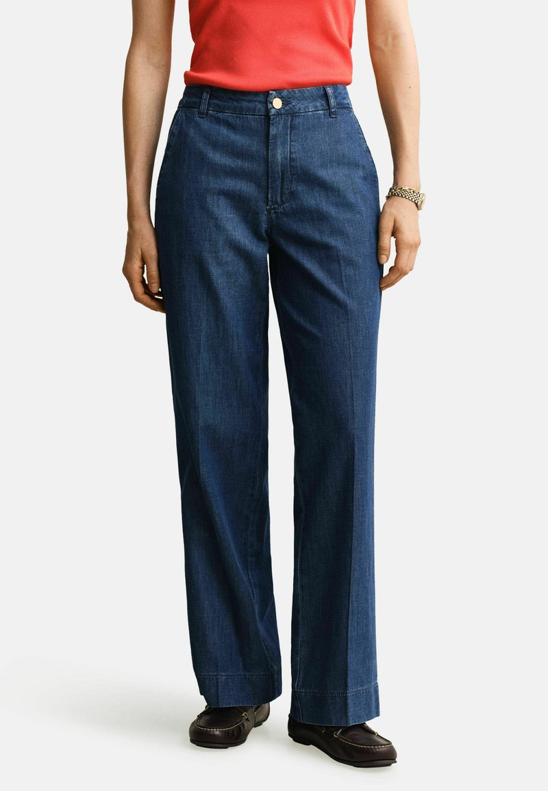 Personne portant un jean large taille haute bleu foncé, un haut rouge, des mocassins marron et une montre en or au poignet gauche.