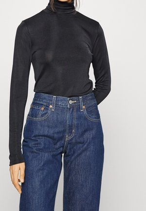 Osoba ubrana w dopasowany czarny golf z długimi rękawami, wpuszczony w wysokowasne niebieskie dżinsy na jednolitym tle.