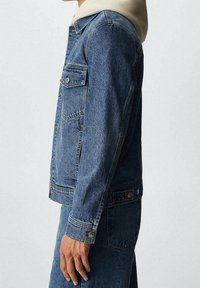 Vista lateral de una persona que lleva una chaqueta de mezclilla azul sobre una sudadera con capucha beige y jeans de mezclilla azul a juego, sobre un fondo liso.