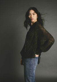 Blouse noire transparente à motif écossais, dotée de manches larges. Associée à un jean bleu taille haute, le modèle se pose contre un fond sombre.