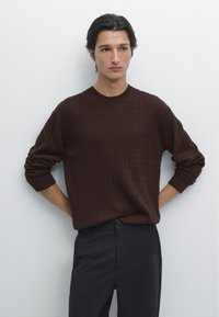 Hnědý pletený svetr s texturovaným vzorem, kulatým výstřihem a žebrovanými manžetami, doplněný tmavými kalhotami na jednobarevném pozadí.