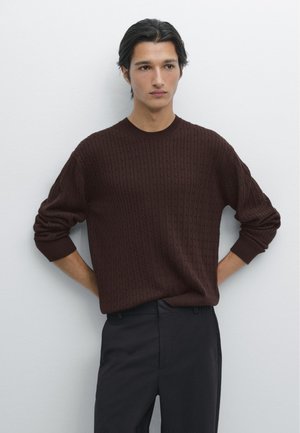 Hnědý pletený svetr s texturovaným vzorem, kulatým výstřihem a žebrovanými manžetami, doplněný tmavými kalhotami na jednobarevném pozadí.