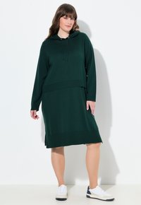 Abito felpa verde a maglia con maniche lunghe, dettagli a costine e tasca a marsupio. Indossato con sneaker bianche e blu, esposto su uno sfondo bianco.