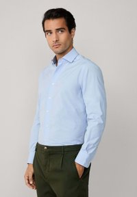 Camisa azul clara de botones con mangas largas, un cuello clásico y tela suave, combinada con pantalones verde oscuro, que cuentan con bolsillos laterales.