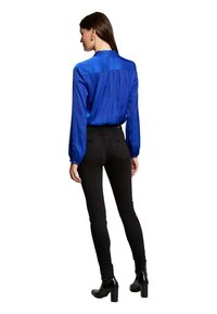 Blusa di raso blu con colletto alto, maniche lunghe a palloncino e pieghe sul retro, abbinata a jeans skinny neri e stivaletti.