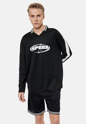 Młody mężczyzna ubrany w czarną koszulkę z długim rękawem z białym logo "SPEED off roading" oraz czarne sportowe spodenki z białymi paskami, stojący na białym tle.