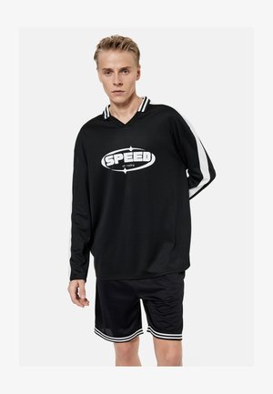Jonge man die een zwart shirt met lange mouwen draagt met een wit "SPEED off roading" logo en zwarte sportshorts met witte strepen, staand tegen een witte achtergrond.