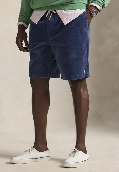 Marineblå kordfløjlsshorts med snøre i taljen, sidelommer og et diskret logo; kombineret med hvide sneakers og en grøn sweater.