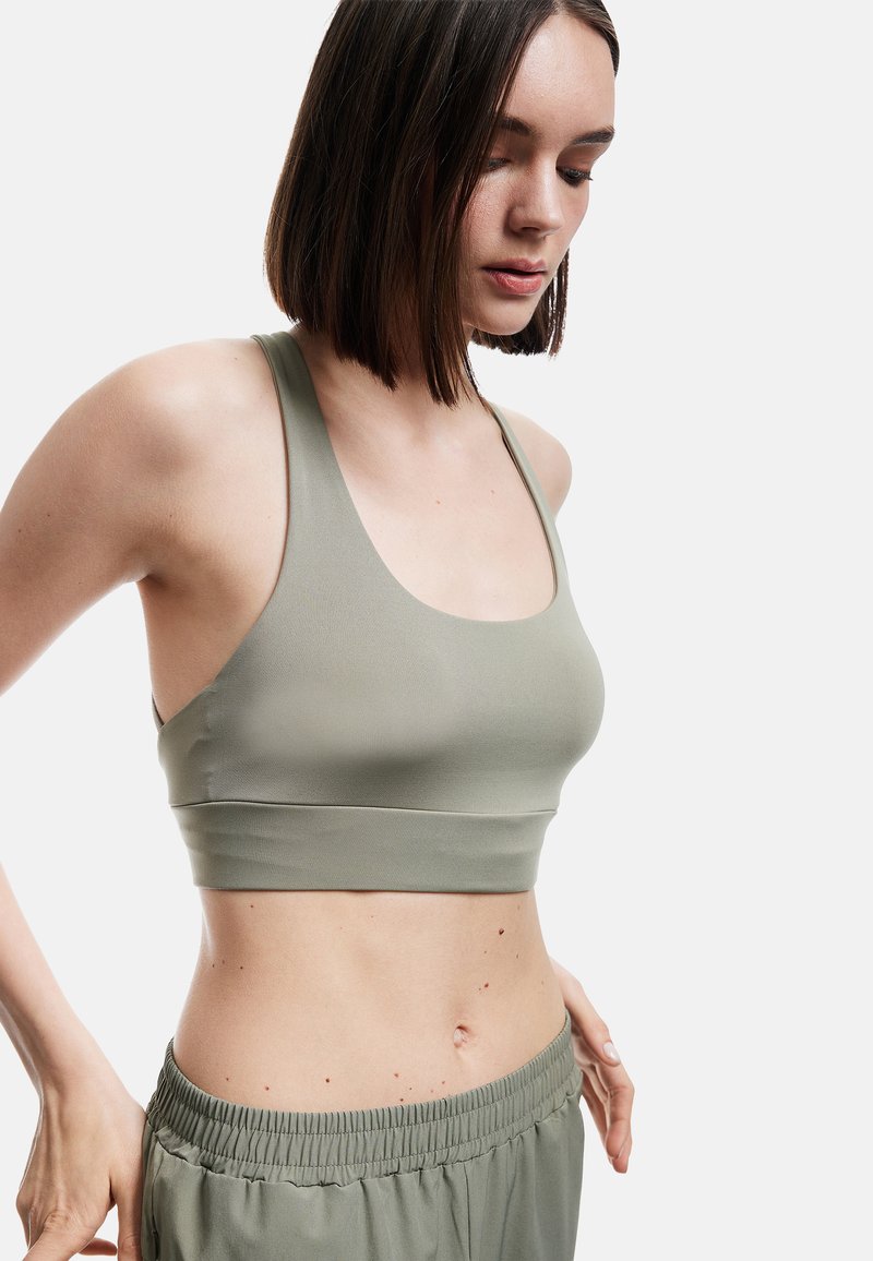 Młoda kobieta poprawiająca oliwkowozielony sportowy biustonosz i dopasowane spodnie z elastycznym pasem, spoglądająca w dół na jednolitym tle.