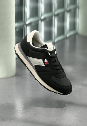 Schwarz-weißer Sneaker mit Schnürdesign und strukturierter Sohle, schwebend vor einem modernen, gekachelten Hintergrund dargestellt.