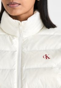 Hvit pufferjakke med høy krage, med horisontale quiltede seksjoner og en liten rød "CK"-logo på venstre side. Glidelås lukking.